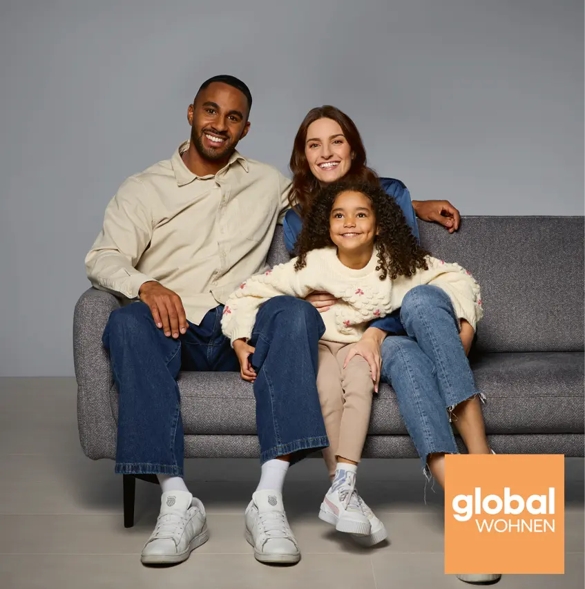 Glückliche Familie sitzt auf grauem Sofa im modernen Wohnzimmer-Stil. Eltern und Kind posieren entspannt für Wohnmöbel-Kampagne von global wohnen. Gemütliche Couch in zeitlosem Design für Familien und Wohnkomfort.